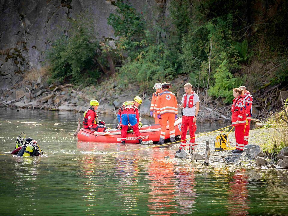 BRK-Retter stehen am Ufer eines Sees, ein Boot hat angelegt ein Taucher ist im Wasser