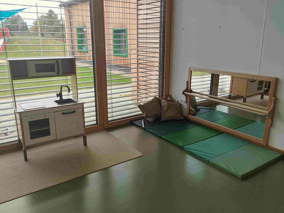 Eine Spielküche steht vor dem Fenster, ein Spiegel mit Haltestange steht an der Wand auf einer Matte